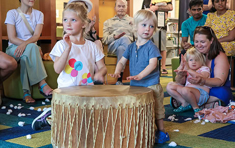 Two young children play a large communal drum while a group of smiling adults and children watch and clap along during a family music event.