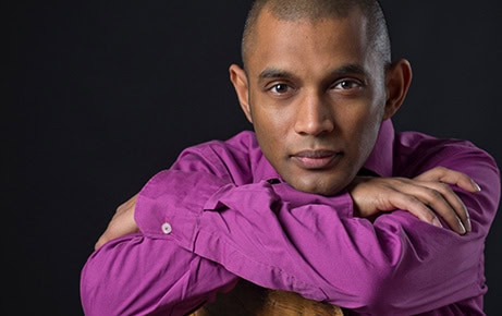 Composer Dinuk Wijeratne rests his arms on the back of a chair and looks warmly at the camera against a dark background.