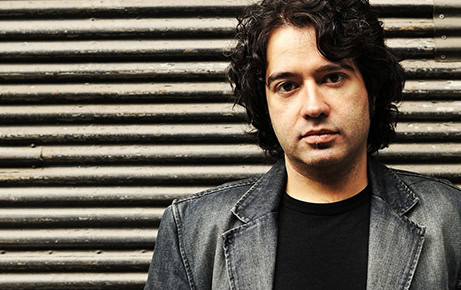 Turkish composer and electronic artist Erdem Helvacioglu stands against a corrugated metal wall, looking directly at the camera.