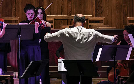 A conductor leads a group of string musicians during a live performance inside a wood-paneled concert hall.