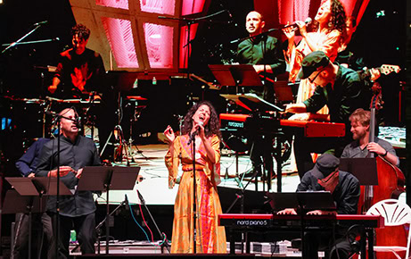 Musicians and singers perform energetically on an outdoor stage at night, with a large video screen behind them projecting their live performance.