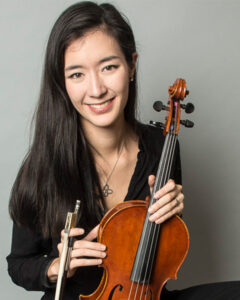 Alexa Brown is holding her viola and smiling, wearing black attire in front of a grey background