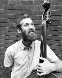 a black and white picture of Sam Beck-Johnson holding a double bass against a brick wall background