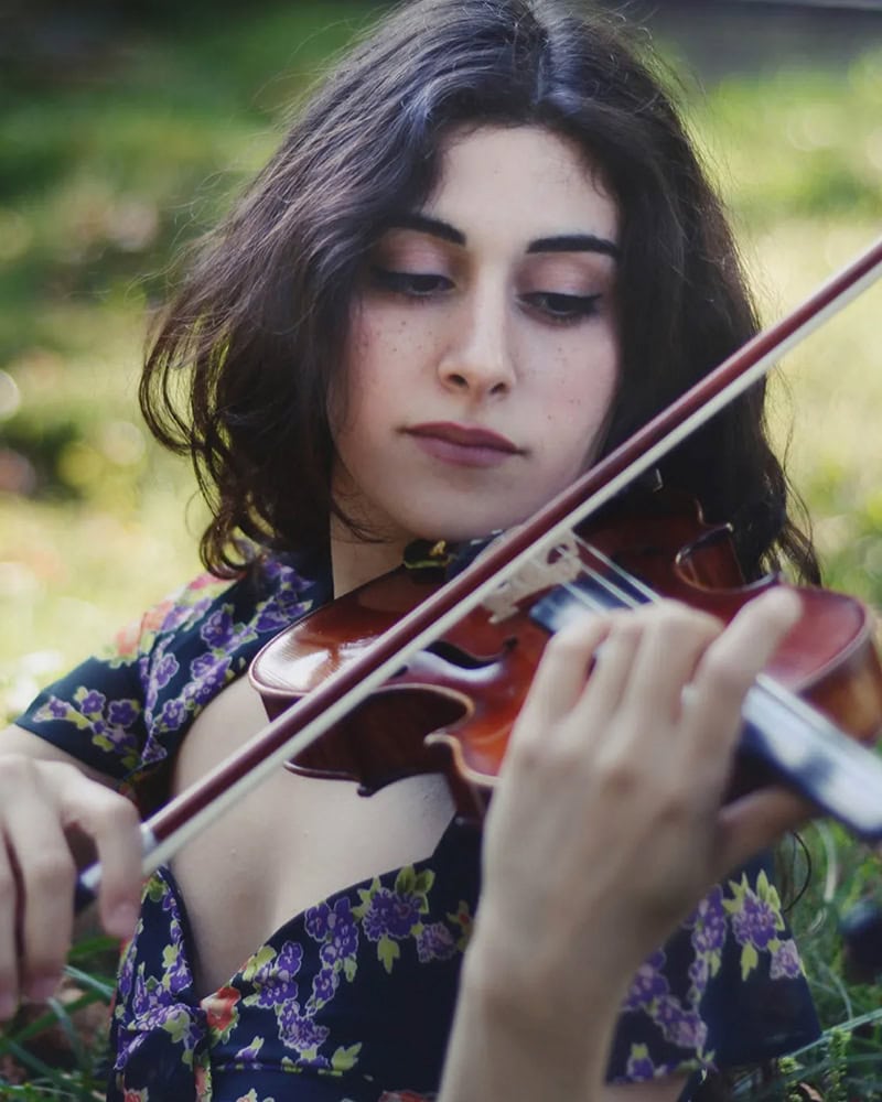 Layale Chaker playing violin outdoors in a floral dress, eyes focused on the instrument