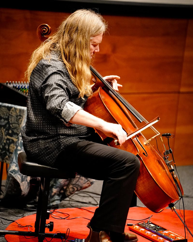David Downing playing his cello on a stage with a red background and electronic pedals at his feet
