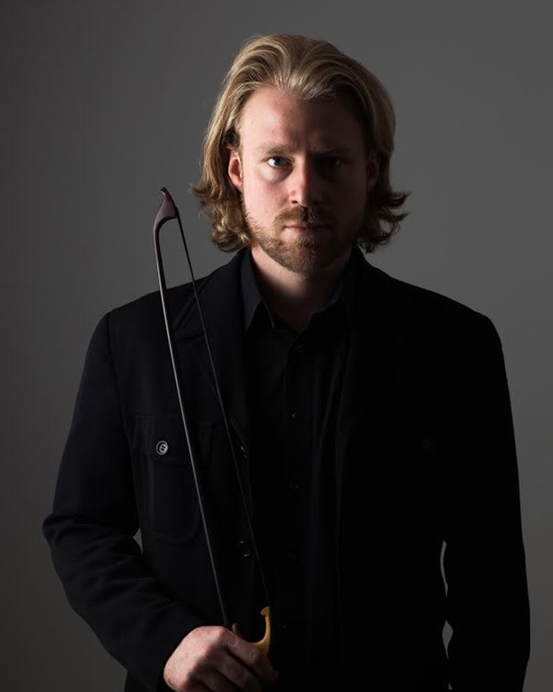 Erik Higgins holding a double bass bow in a black coat, standing against a gray background