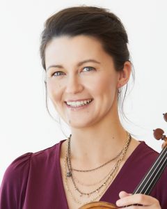 Smiling violinist holding instrument, wearing a burgundy top and layered necklaces against a white background