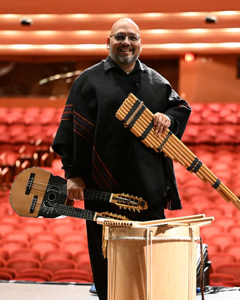 Oscar Rios Pohirieth on stage holding Andean instruments including zampoñas, charango, and bombo, in a concert hall