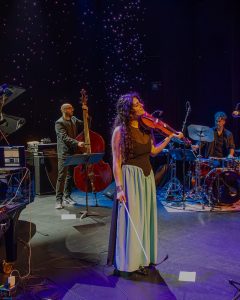 Sarafand ensemble performing live on stage, with Layale Chaker on violin and band members on bass, percussion, and keyboard