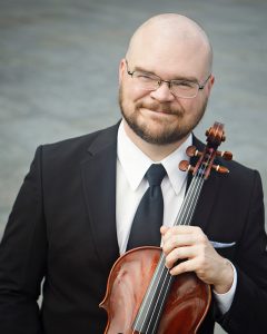 Violist Tyler Sieh wearing a black suit and tie, holding his viola and smiling at the camera