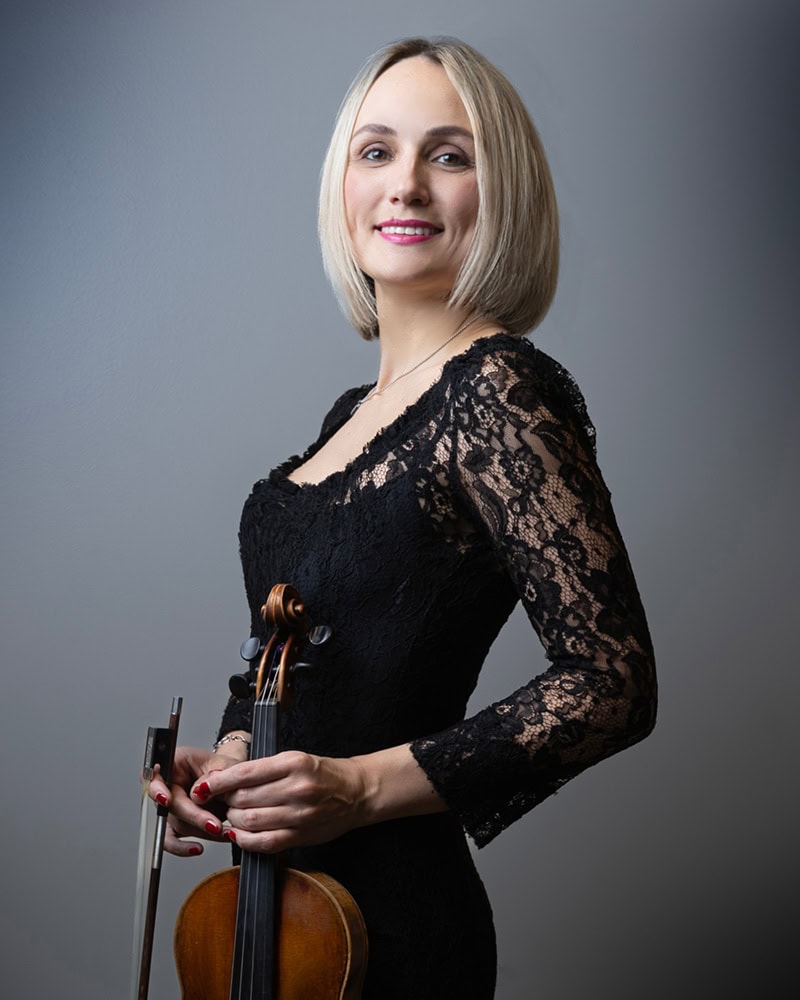 Violinist Olga Smola in a black lace dress holding a violin and bow, smiling in front of a grey background