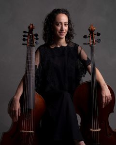 Baroque cellist Anna Steinhoff seated between a viola da gamba and a cello, wearing black and smiling softly