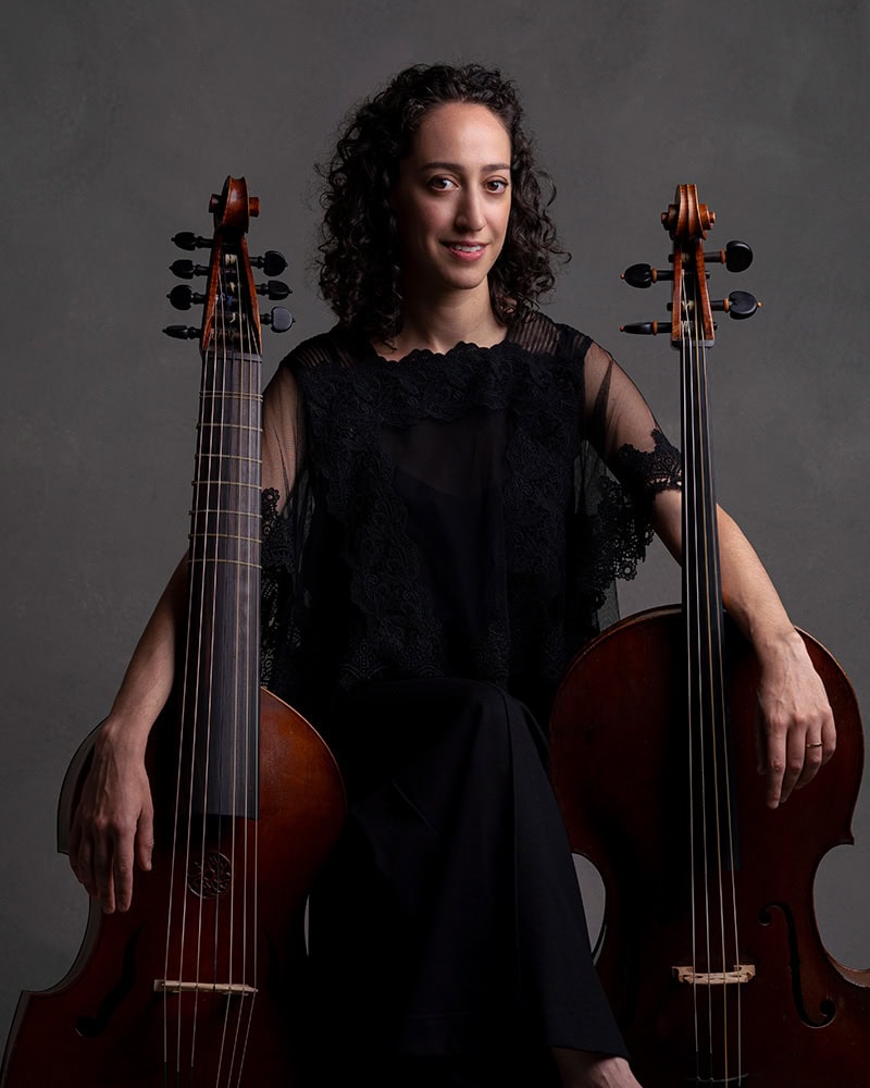 Baroque cellist Anna Steinhoff seated between a viola da gamba and a cello, wearing black and smiling softly