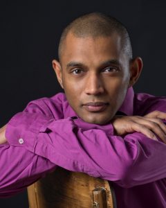 Dinuk Wijeratne in a magenta shirt leaning on a chair and looking into the camera