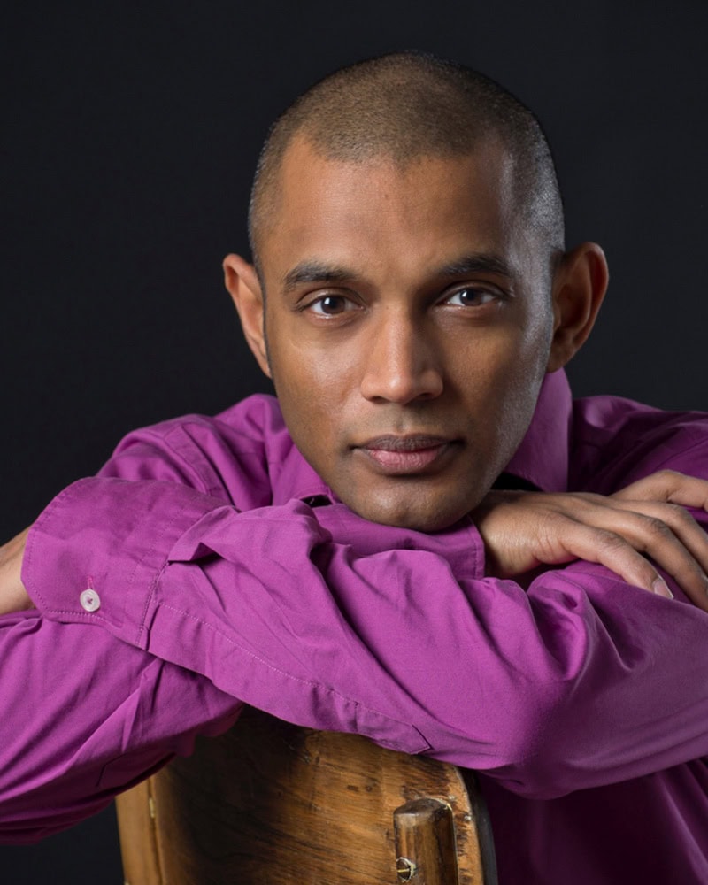 Dinuk Wijeratne in a magenta shirt leaning on a chair and looking into the camera