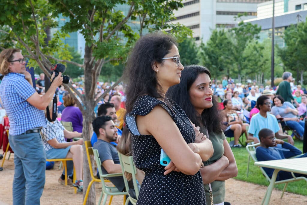 Two women standing, looking towards the stage with a large seated audience in the background.