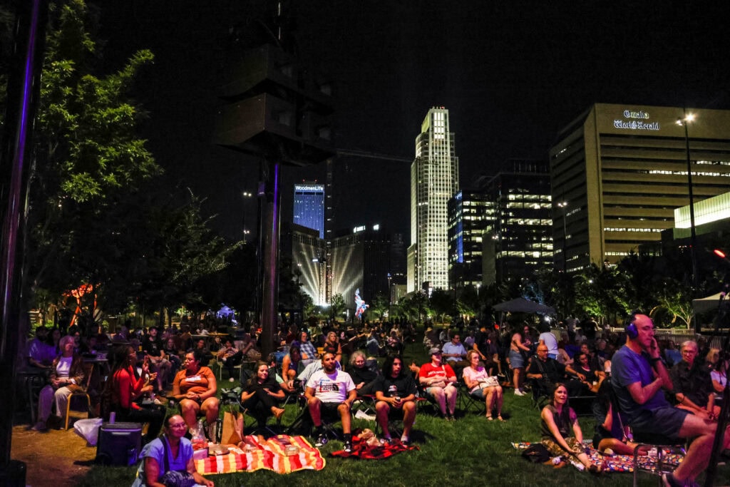 Audience members sit on the lawn of the Gene Leahy Mall at dusk, with the Omaha skyline in the background