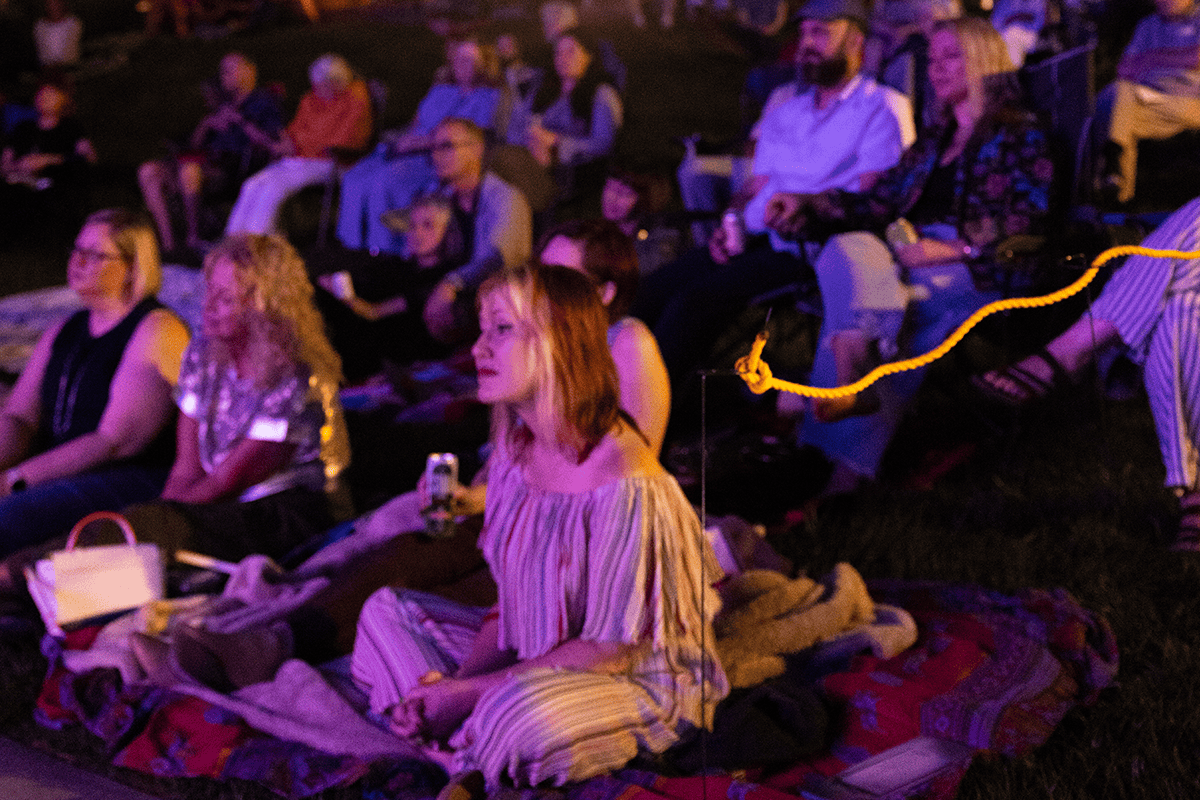Woman sitting on a blanket enjoying the music at an outdoor concert within a large crowd