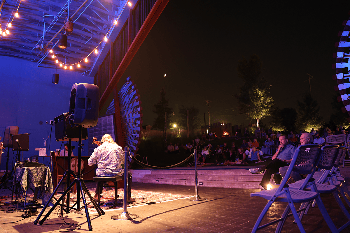 A view from backstage with a cellist performing in a semi-outdoor space with speakers and lighting.