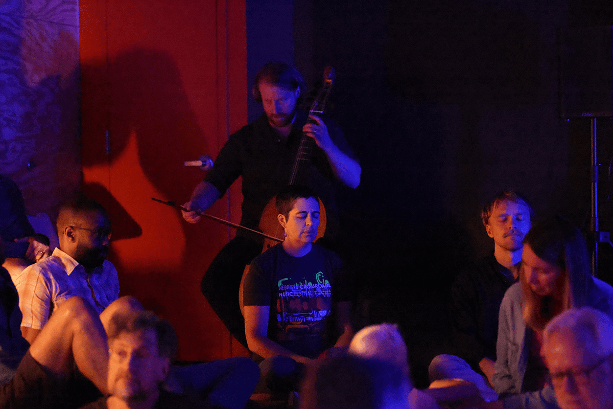 Erik Higgins performs on viola da gamba during a soundscape performance with dark lighting and people sitting on the floor.