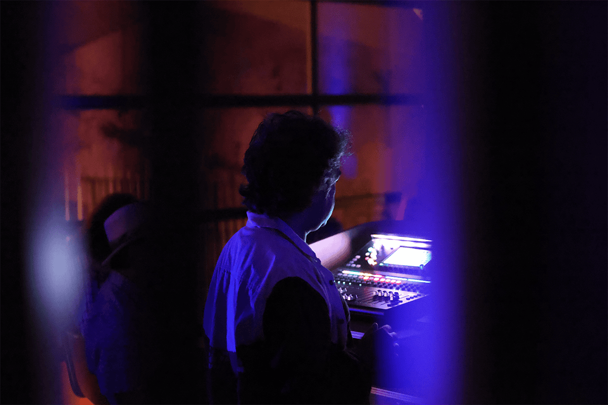 Erdem Helvacioglu sitting at his mixing station, framed through a grate and in profile, illuminated by the mixing board.
