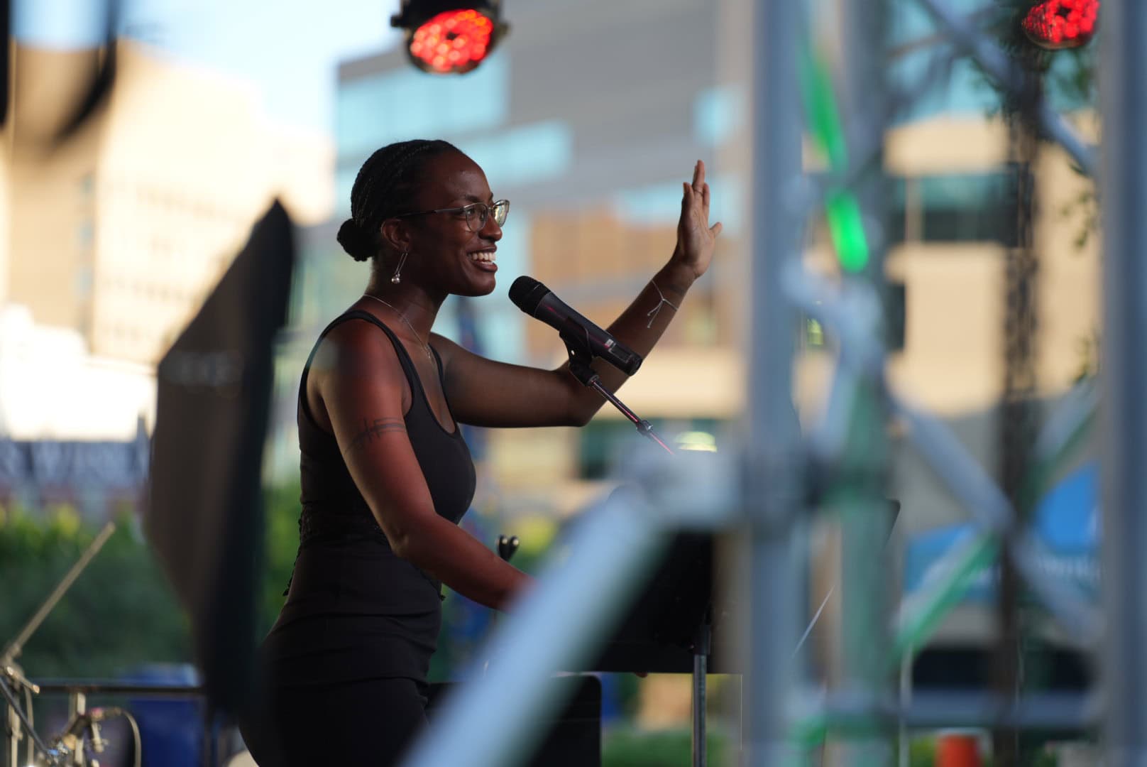 Nebraska State Poet Jewel Rodgers speaking outdoors with arm outstretched on a stage with red lighting with Lincoln's blurred skyline in the background