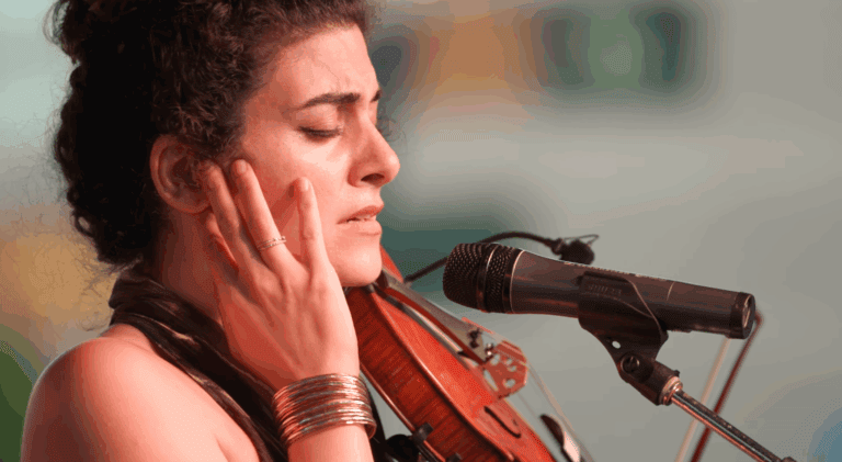 Layale Chaker sings into a microphone while holding a violin.
