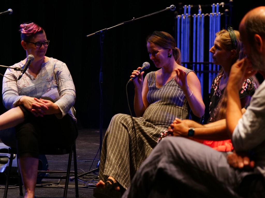 Four people speaking into microphones on a darkly lit stage.